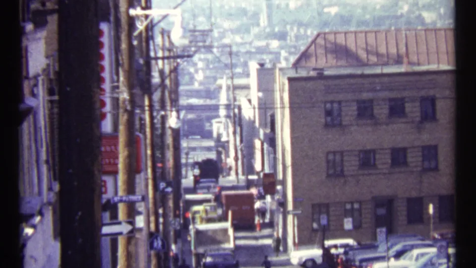 1967 Ottawa street frame with cars, pedestrians, low-rise buildings, utility poles, wires, and a hazy distant hillside beyond the block.