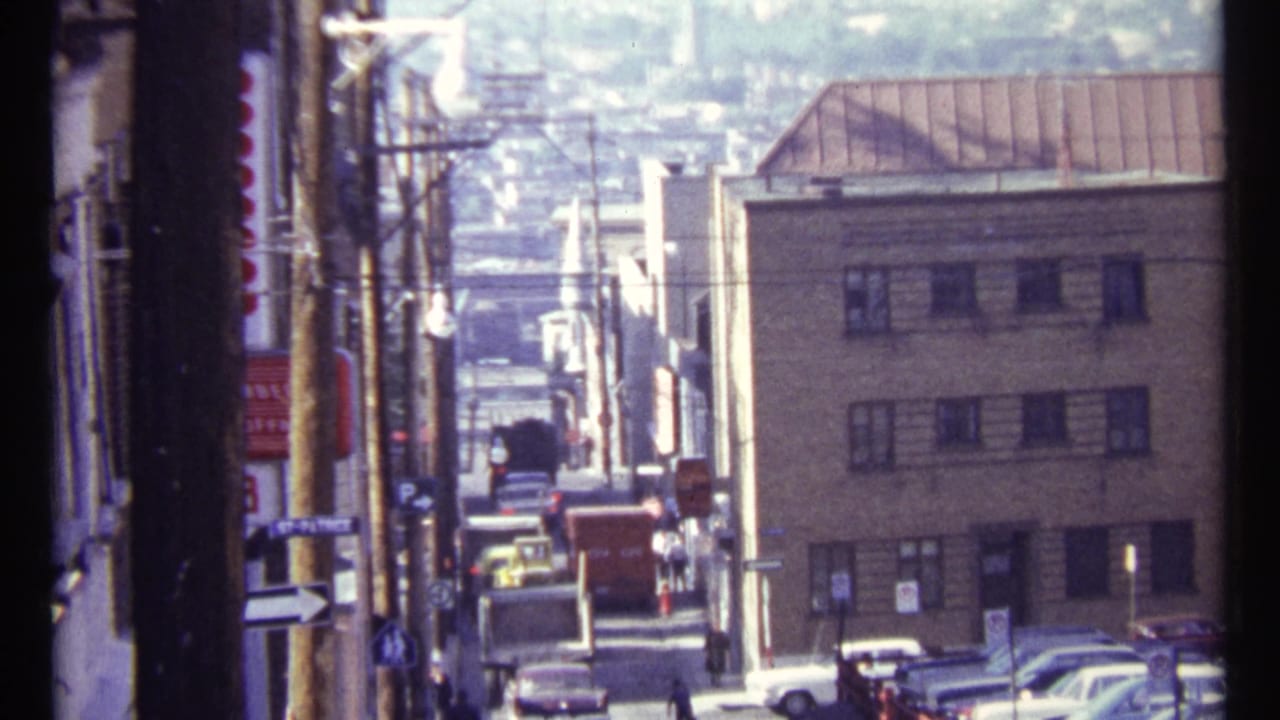 Narrow city street viewed downhill with older cars, utility poles, and low buildings.