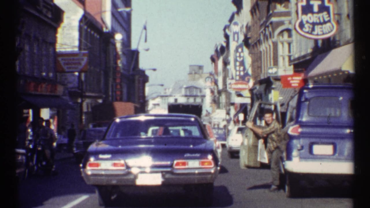 Busy street with a blue sedan, storefront signs, parked vehicles, and pedestrians in bright daylight.