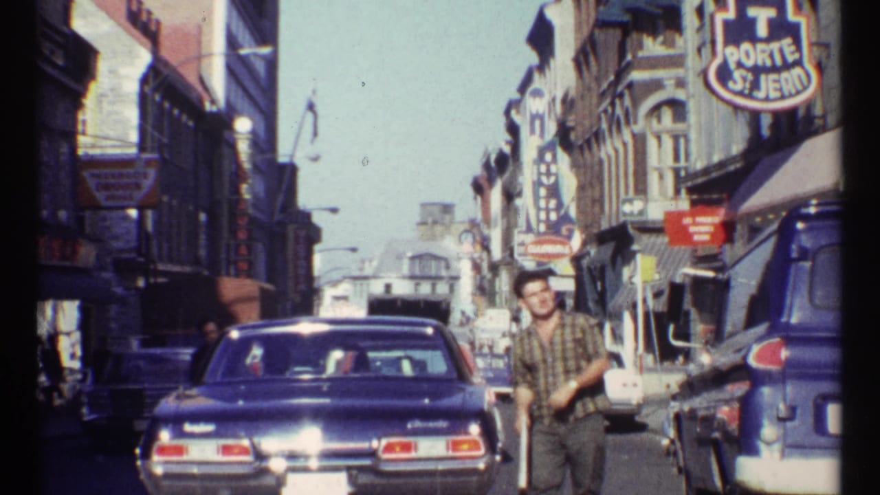 A man in a plaid shirt passes near a blue sedan and blue van on an Ottawa commercial street in 1967 archival home-movie footage.