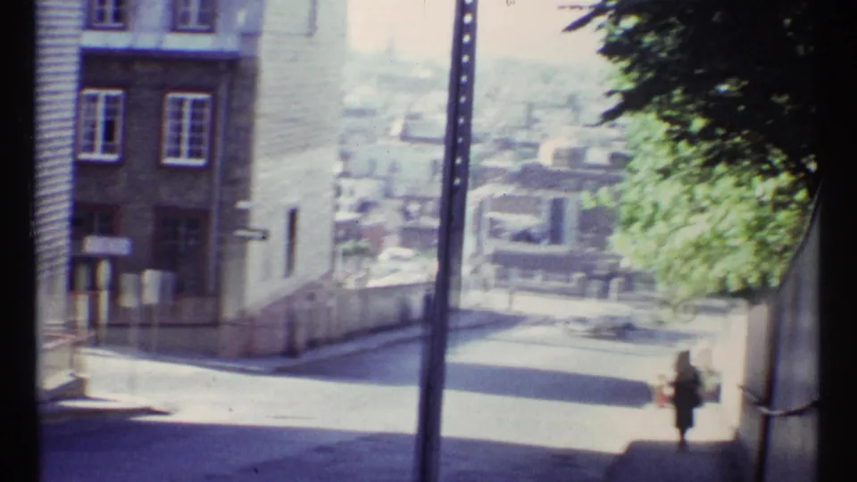 A sloped Ottawa street appears in soft 8mm footage, with older buildings, a utility pole, trees, and a distant pedestrian under bright haze.