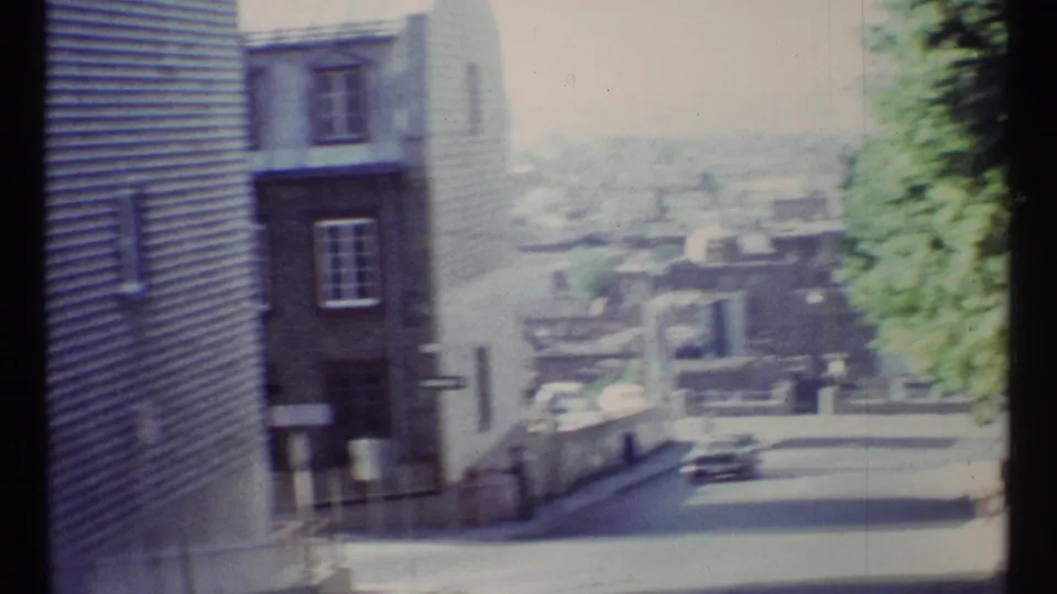 Soft 1967 Ottawa street view looking downhill past older buildings and trees, with distant cars and bright exposure from the 8mm film scan.