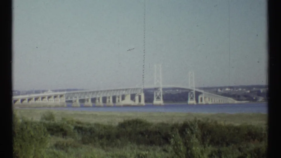 1967 Ottawa home-movie view of a pale bridge spanning water, seen from a grassy shoreline under open sky.