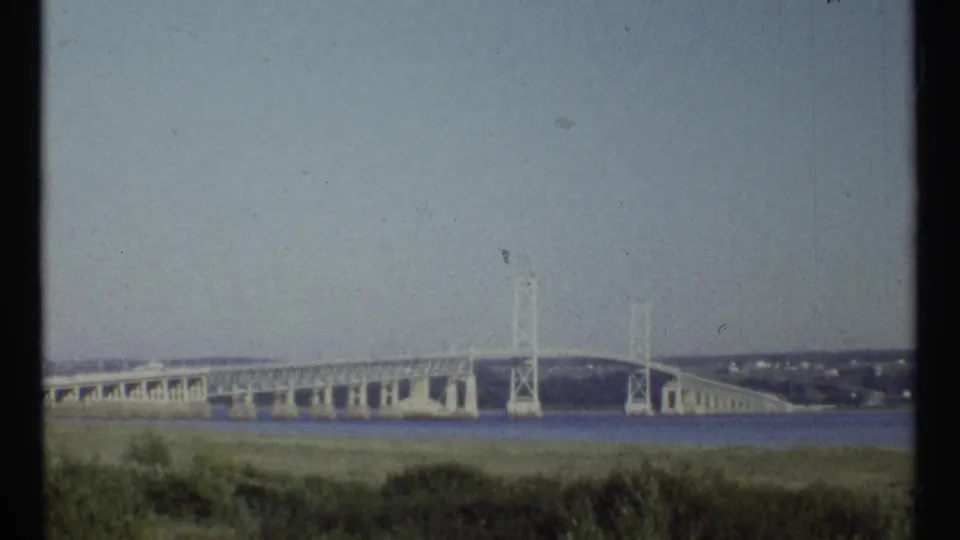 Soft 8mm frame of the Ottawa bridge scene, showing the long span, towers, water, and low shoreline vegetation.