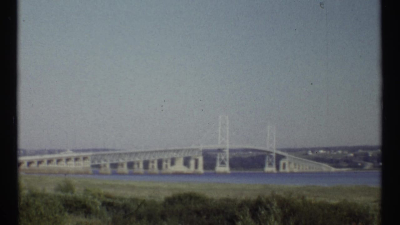 Distant bridge or dam over water with grassy foreground and pale sky.