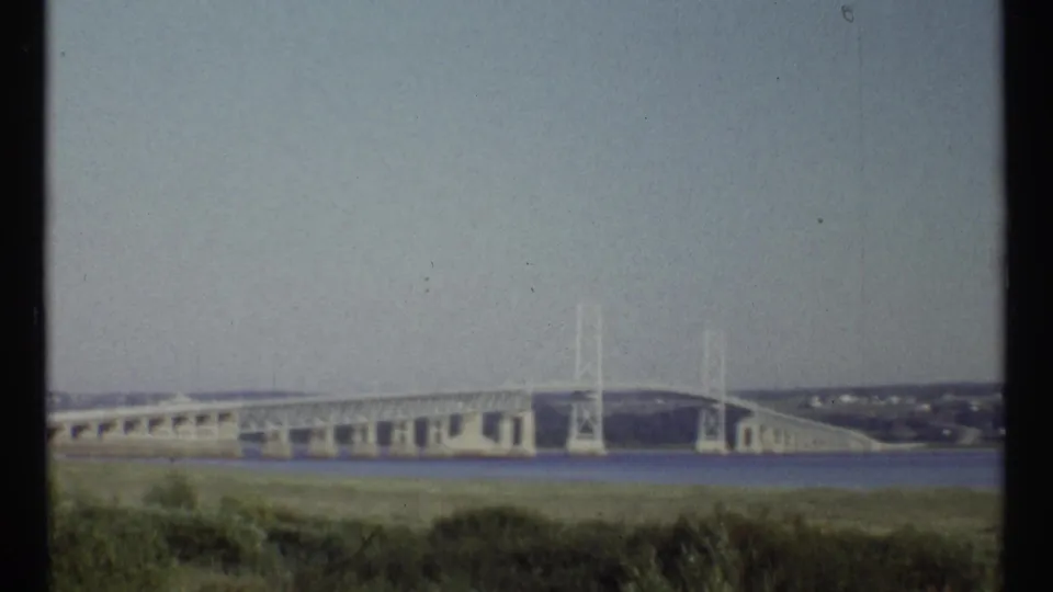 1967 8mm frame from Ottawa showing a distant bridge or dam across water, viewed over grass under a pale sky.