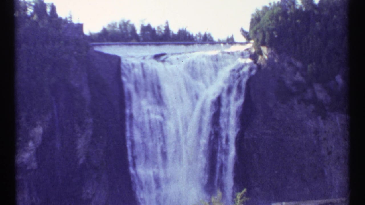 Wide waterfall dropping over a cliff between dark rock walls and trees.