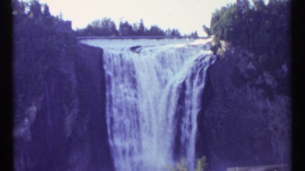 Waterfall pouring over a wide ledge with trees and dark cliff faces around it.