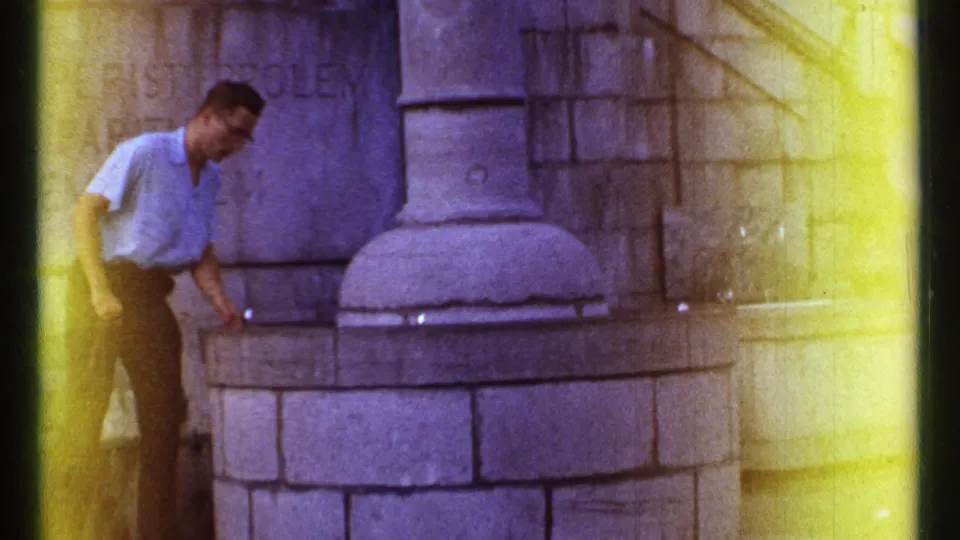 1967 Ottawa frame showing an adult man in a blue shirt and dark trousers beside a stone fountain with Latin lettering behind him.