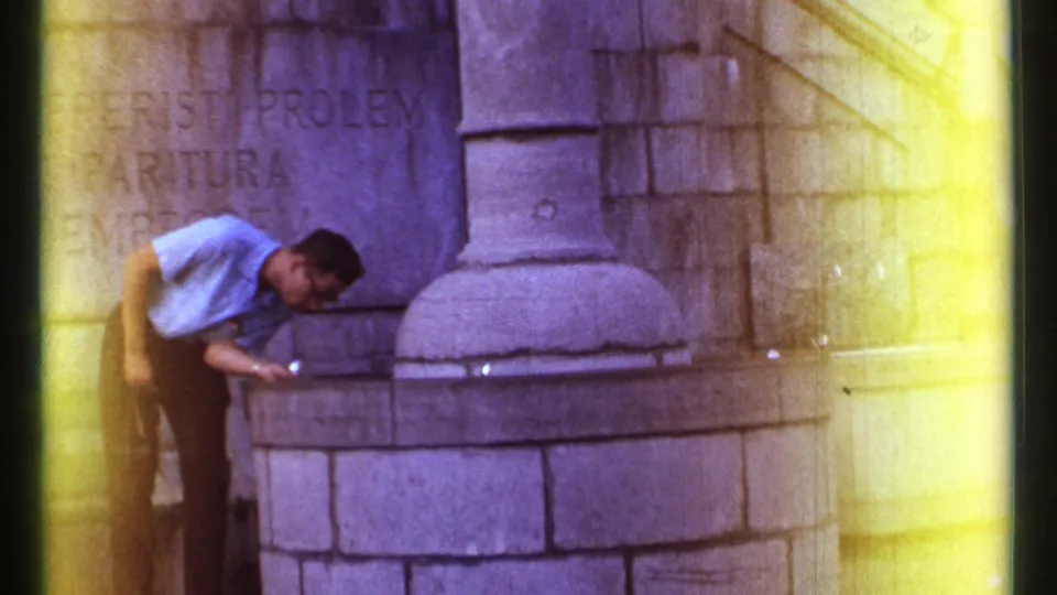 1967 Ottawa home-movie frame of an adult man bending at a stone fountain, with Latin inscription and soft 8mm color flares visible.