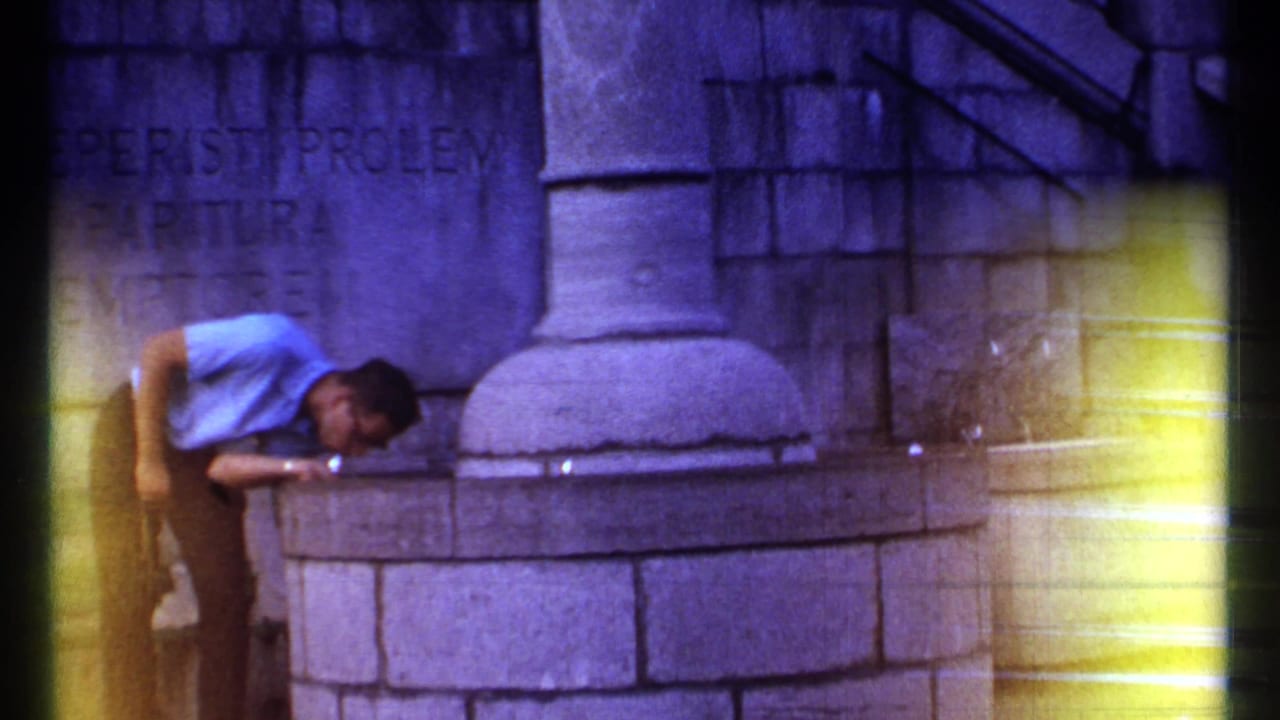 Man bends over a stone fountain beside an engraved wall and column.