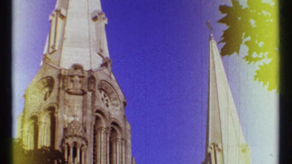 Handheld 1967 Ottawa footage frames two pale cathedral spires with tree leaves and visible color-shifted film texture.