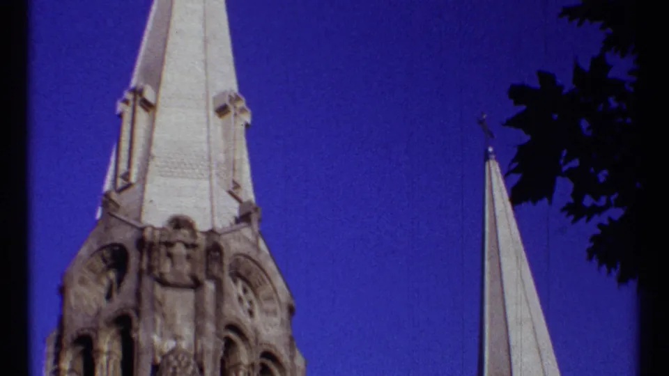 1967 Ottawa home-movie frame showing a pale church steeple, ornate stone tower, smaller spire, and dark leaves at the edge.
