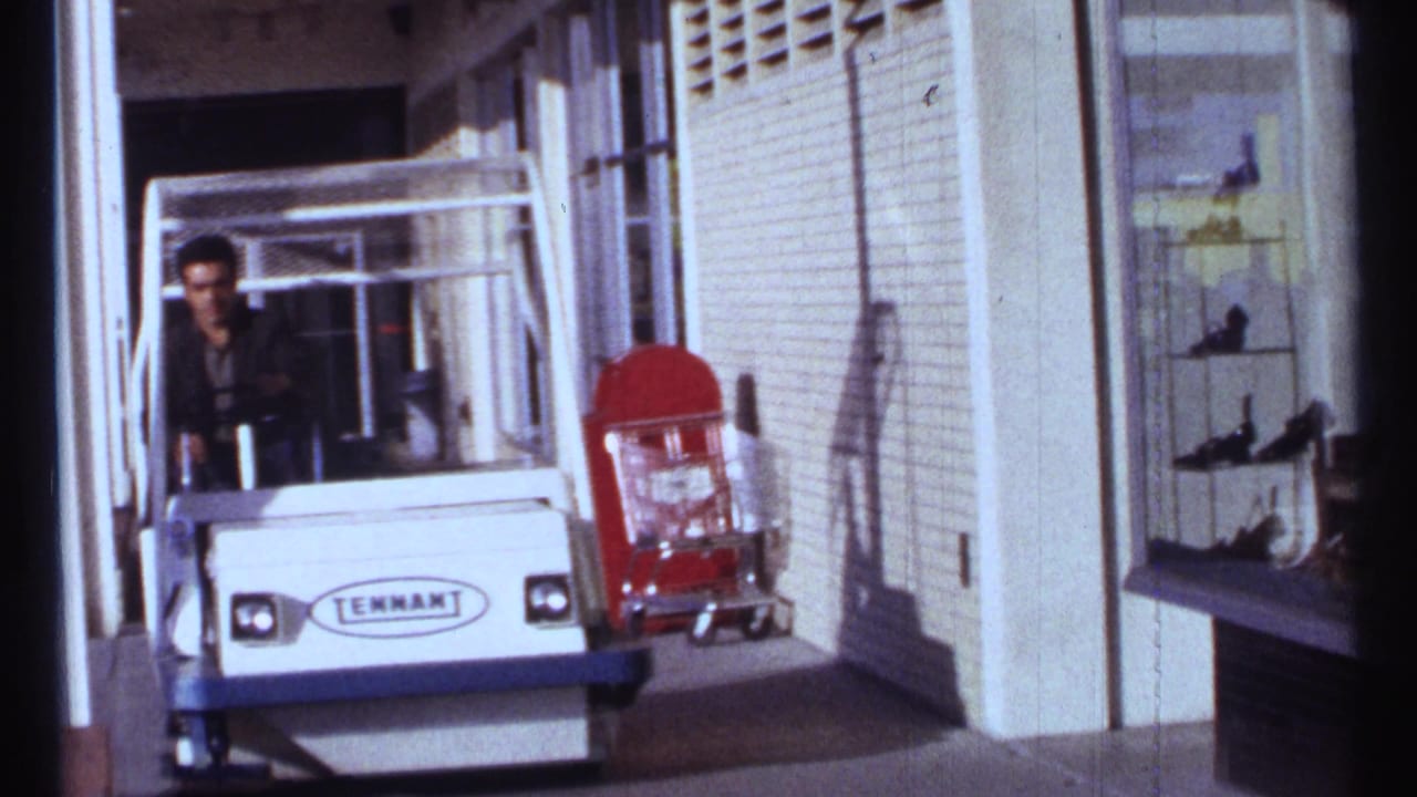 Man driving a white Tennant cart along a storefront sidewalk with a red cart nearby.