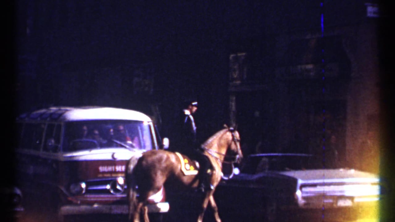 Mounted officer on a horse near period cars and a red and white van on a dim city street.