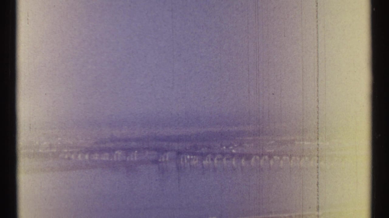 Hazy aerial view across a wide river toward a distant city skyline and long bridge.