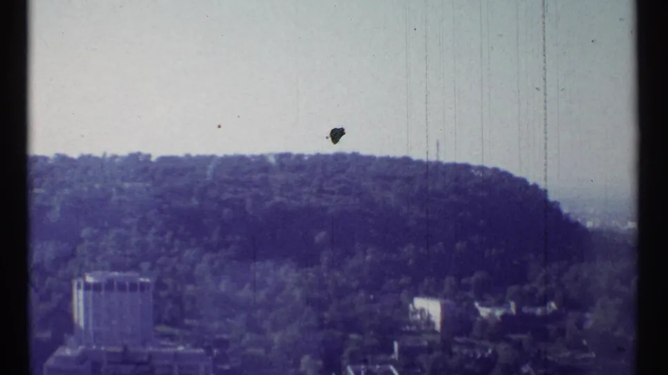 1967 Ottawa view shifting toward a wooded hillside, a rectangular tower, low buildings, and scratched 8mm film texture.