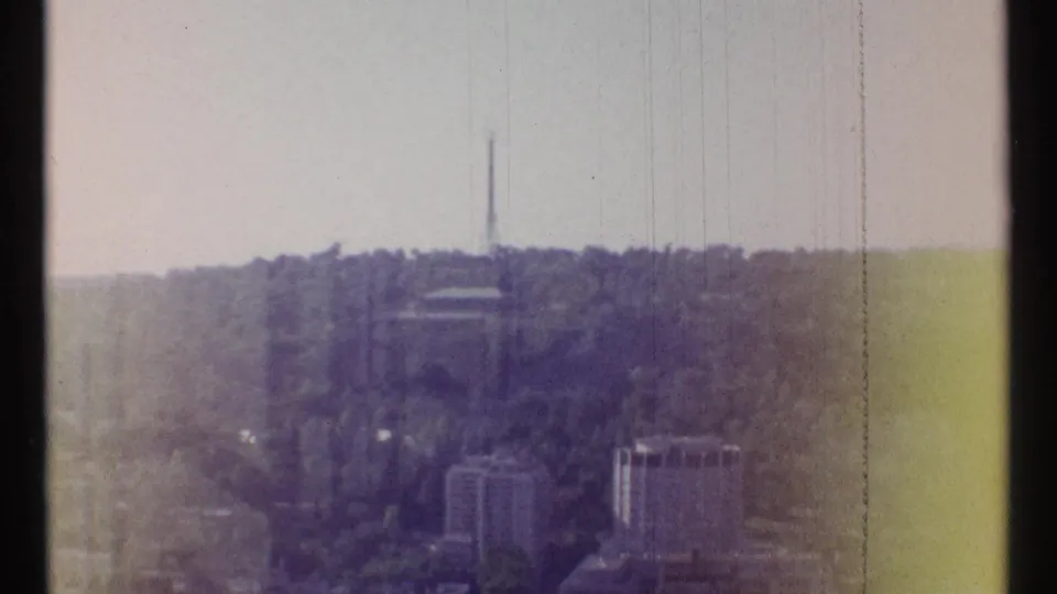 1967 Ottawa frame showing distant buildings below a tree-covered ridge, with a slender tower on the skyline.