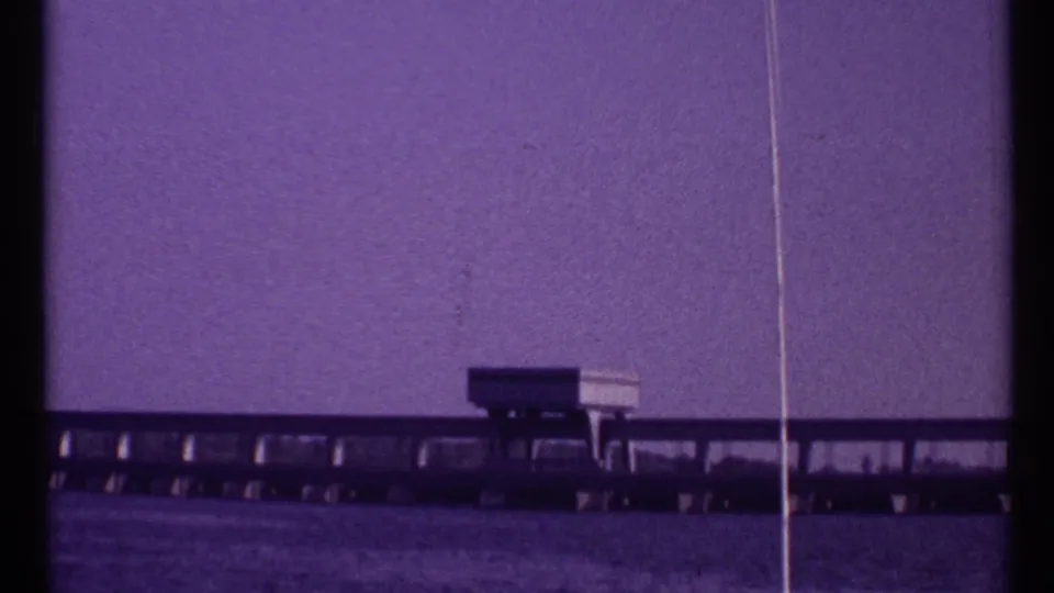 Toronto 1967 frame showing a small building or control station on a low bridge over water, seen through purple-shifted 8mm film.