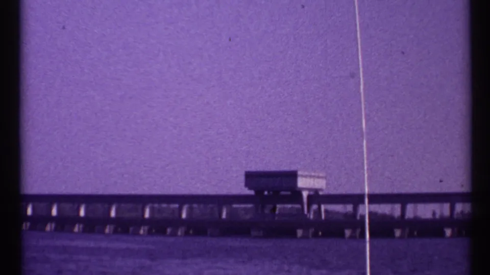 1967 Toronto 8mm frame showing a long low waterfront structure with supports beneath a broad purple-tinted sky.