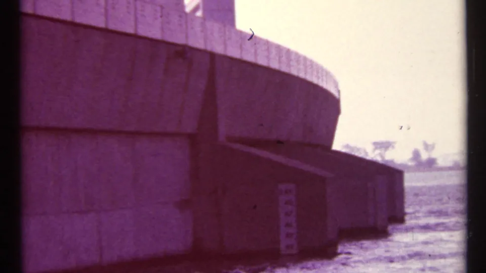 1967 Toronto home-movie frame showing the curved concrete face of the structure beside water, with a distant low shoreline.