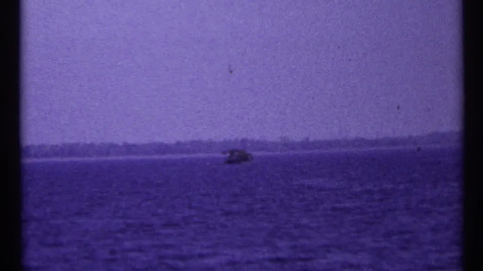 Grainy 1967 Toronto-area home-movie frame of a small boat on dark lake water below a pale, hazy sky.