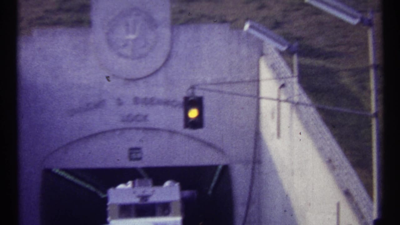 1967 Canadian 8mm frame with an amber signal lit above a white vehicle entering or waiting at a concrete hydroelectric station portal.