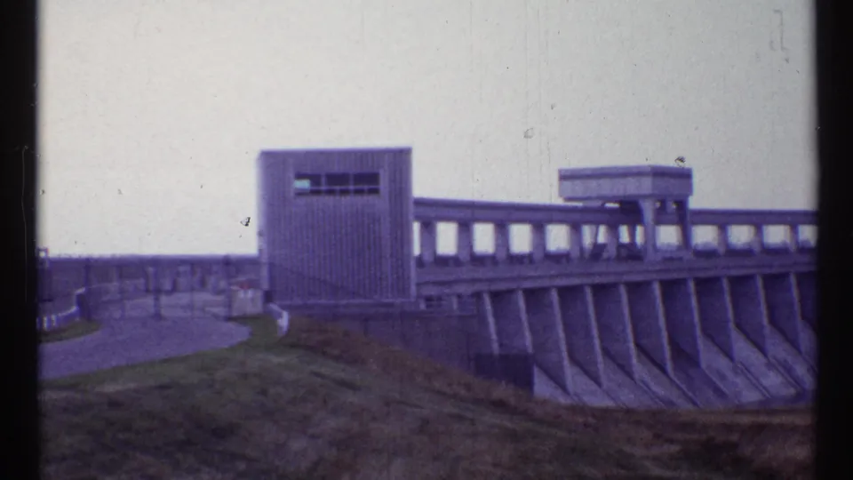1967 Canadian 8mm frame showing a hydroelectric dam spillway, railings, and rectangular control structures from a visitor viewpoint.