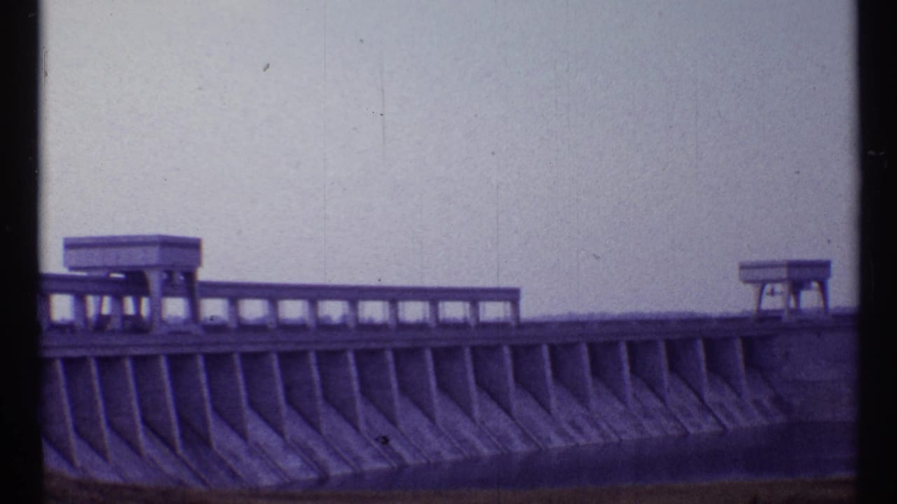 Concrete dam face with railing and two small platform structures under a pale sky.