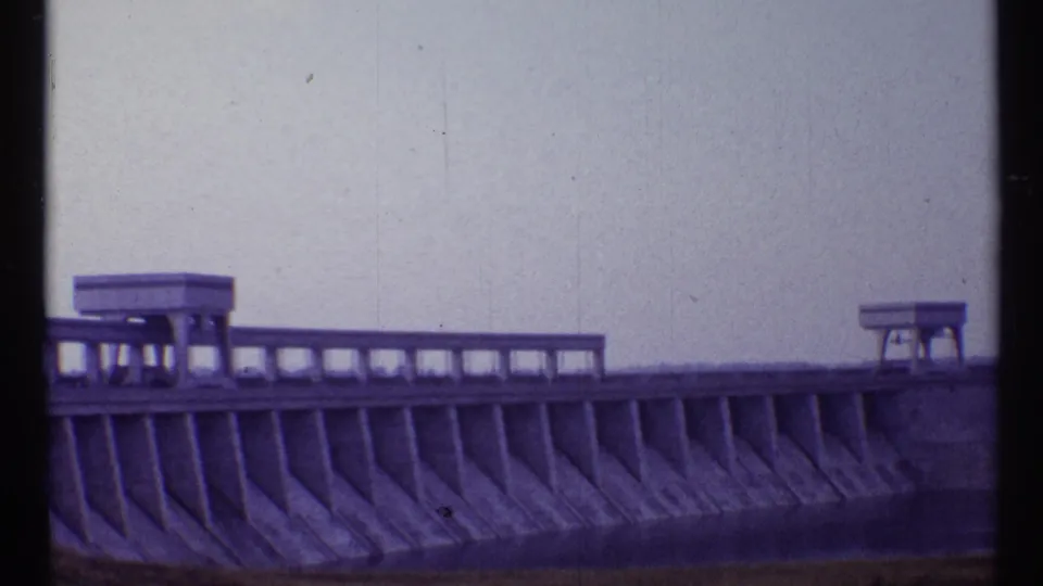 1967 Toronto home-movie frame showing a concrete dam with angled spillway supports, top railing, and small platform structures.