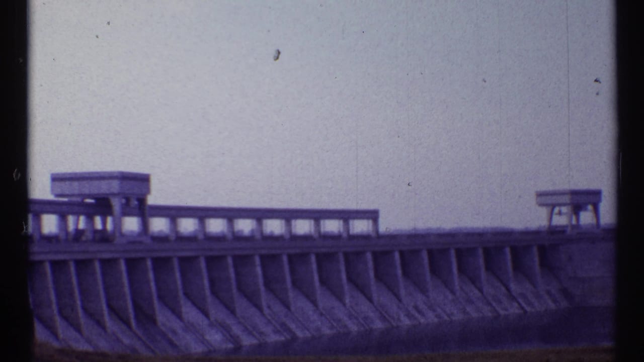 Wide view of a concrete dam section with railings, platform structures, and pale sky.