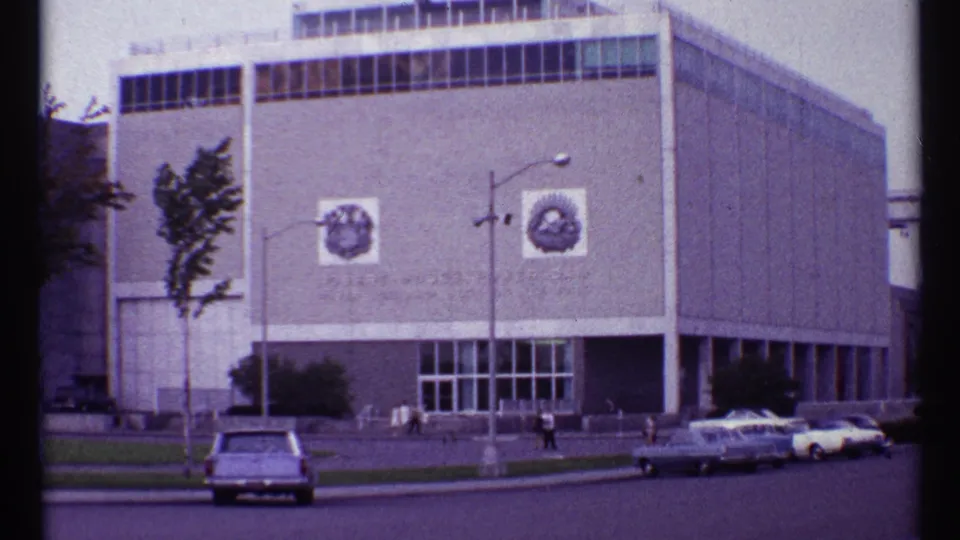 1967 Toronto frame showing a modernist building exterior, wall emblems, streetlamps, small distant people, trees, and vintage cars.