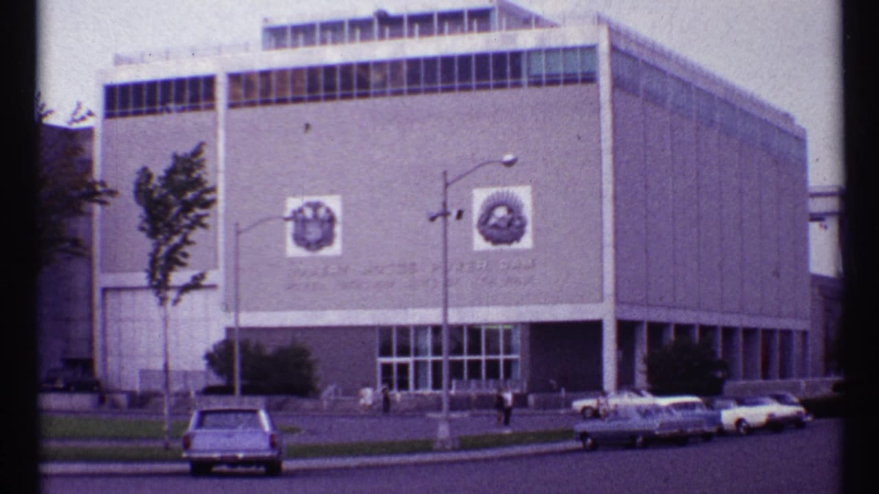 Modernist building with facade emblems, parked cars, streetlamp, trees, and pedestrians.