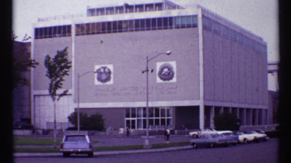 1967 Toronto home-movie view of a modernist building with two facade emblems, parked cars, streetlamp, trees, and pedestrians.