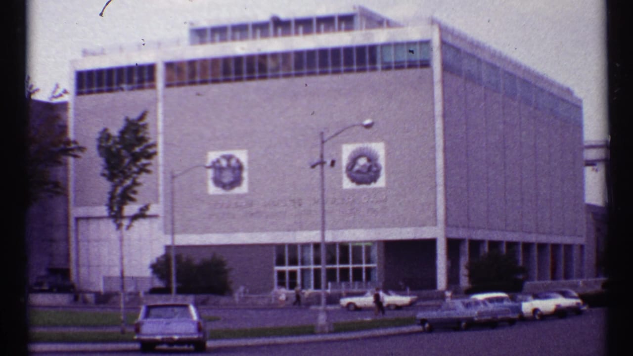 Street view of the same large building with emblems, parked cars, and people near the entrance.