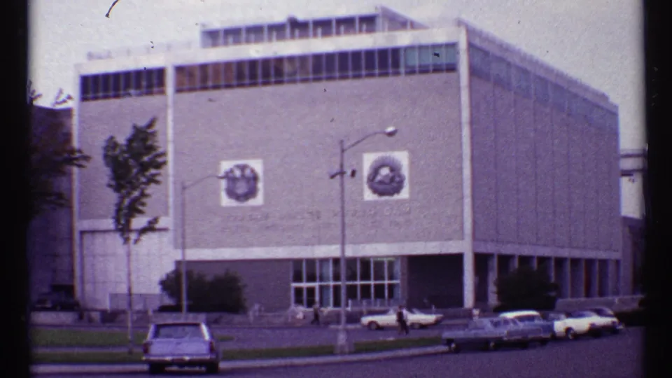 1967 Toronto street view showing a large modernist building, visible facade emblems, parked cars, and pedestrians near the entrance.