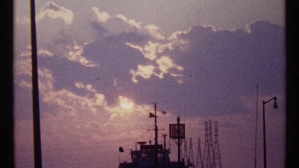 1967 Toronto harbor frame showing the sun breaking through clouds above silhouetted ship structures, masts, poles, and distant industrial forms.