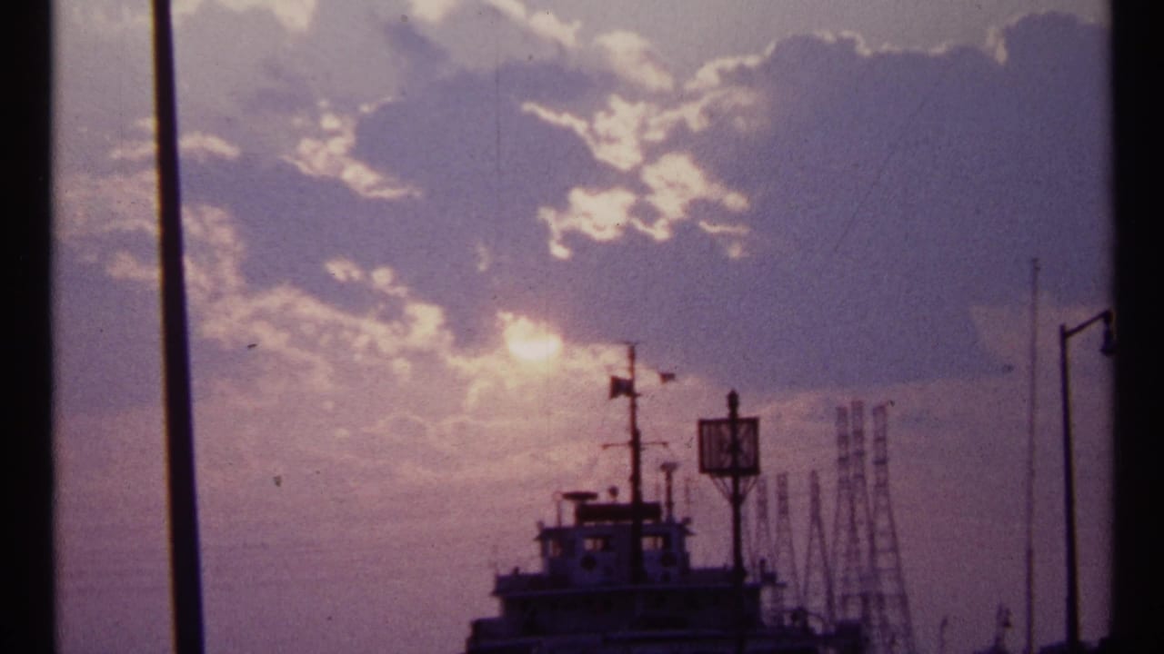1967 Toronto waterfront view showing a bright sun through clouds, a silhouetted vessel, mast, and industrial forms in the distance.