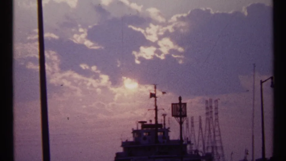 1967 Toronto waterfront view showing a bright sun through clouds, a silhouetted vessel, mast, and industrial forms in the distance.