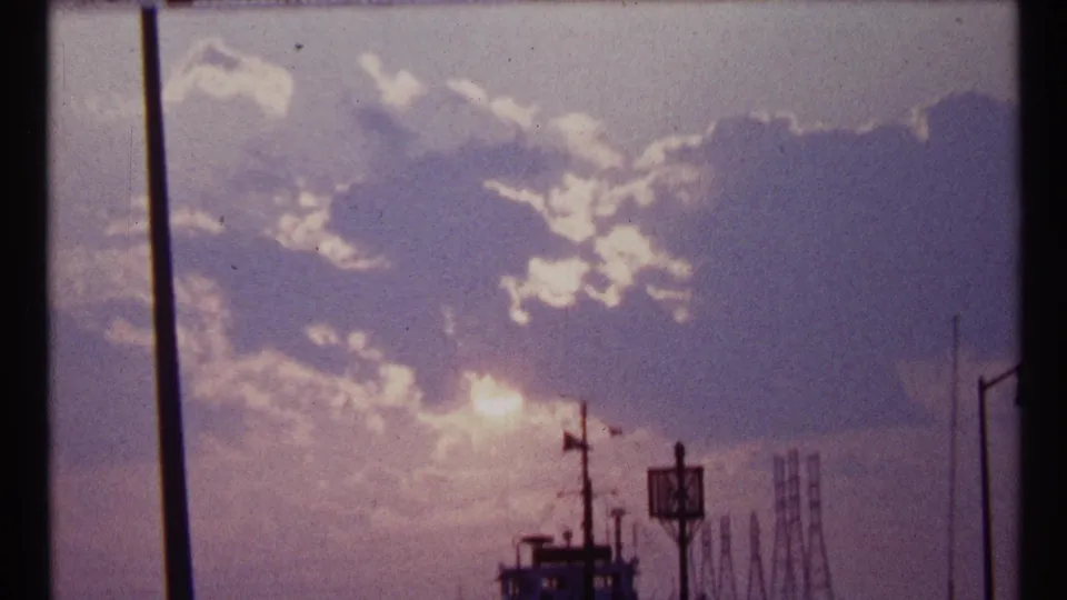 Toronto 1967 home-movie frame with blooming sunlight, purple-tinted clouds, a dark vessel, and faint harbor infrastructure.