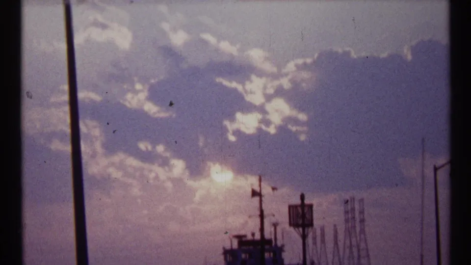 Final 1967 Toronto 8mm frame with layered clouds, strong sun glare, a silhouetted ship, and distant industrial waterfront shapes.