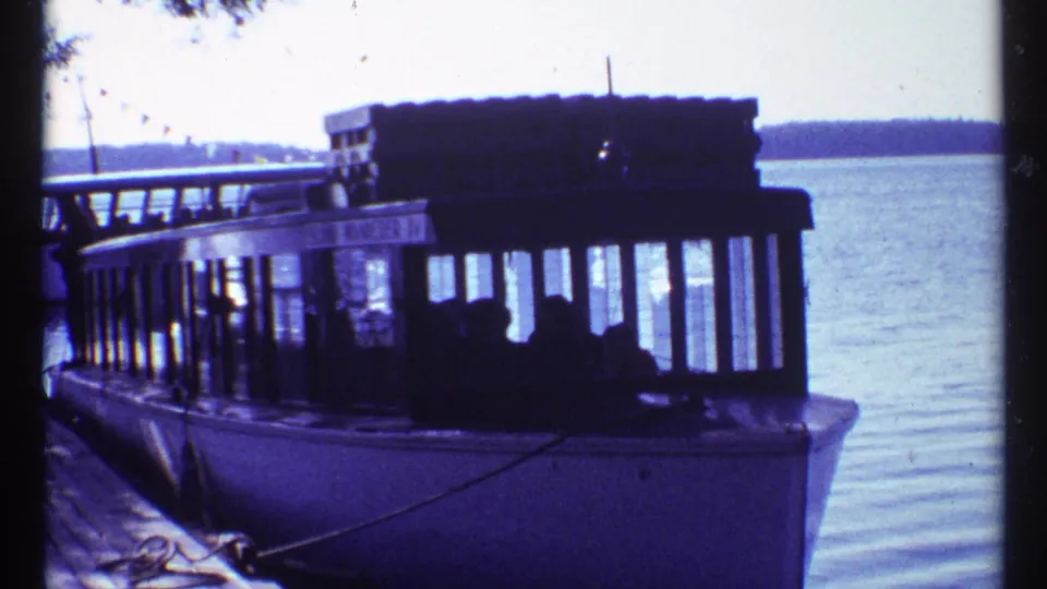 The ferry sits docked at a pier on the Toronto waterfront, with passenger silhouettes inside the cabin and calm water behind it.
