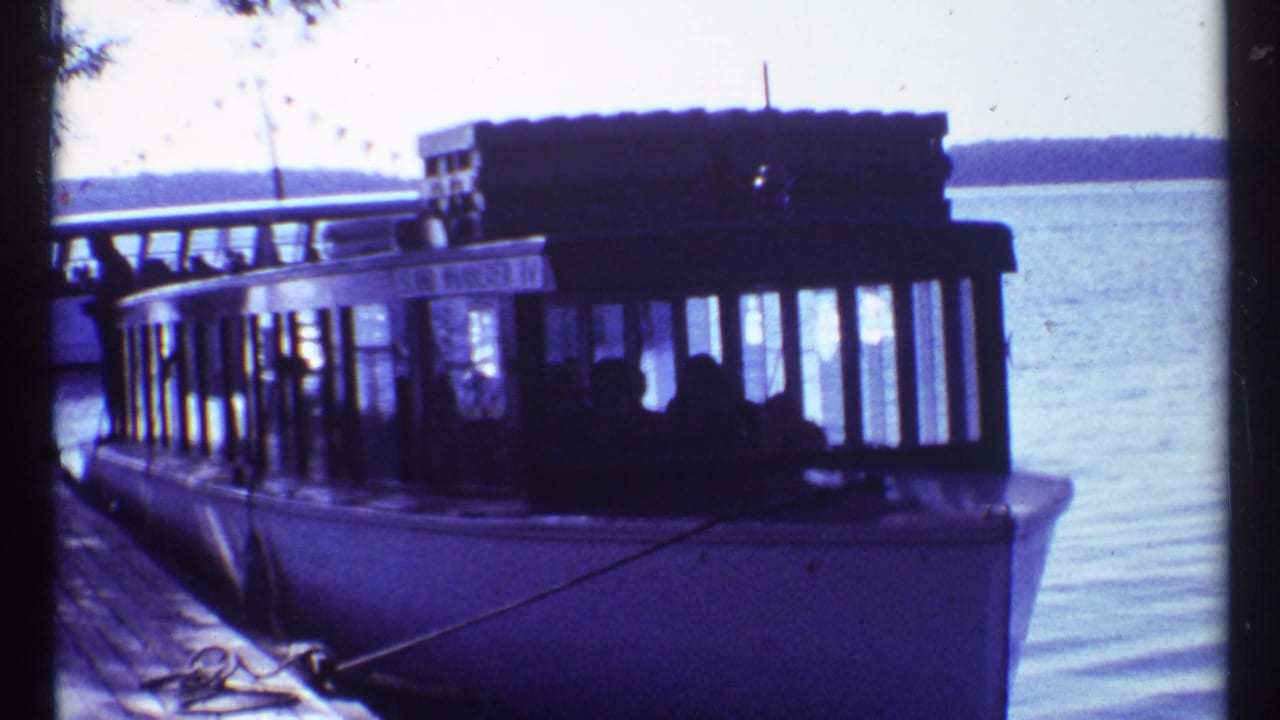 1967 Toronto waterfront home-movie frame showing a docked passenger tour boat, pier edge, lake water, and figures inside the cabin.