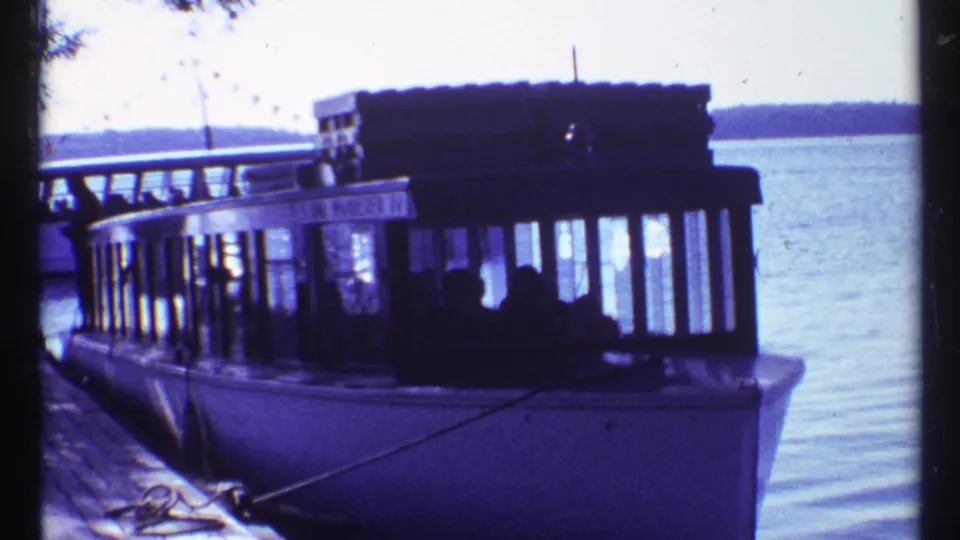 1967 Toronto waterfront home-movie frame showing a docked passenger tour boat, pier edge, lake water, and figures inside the cabin.