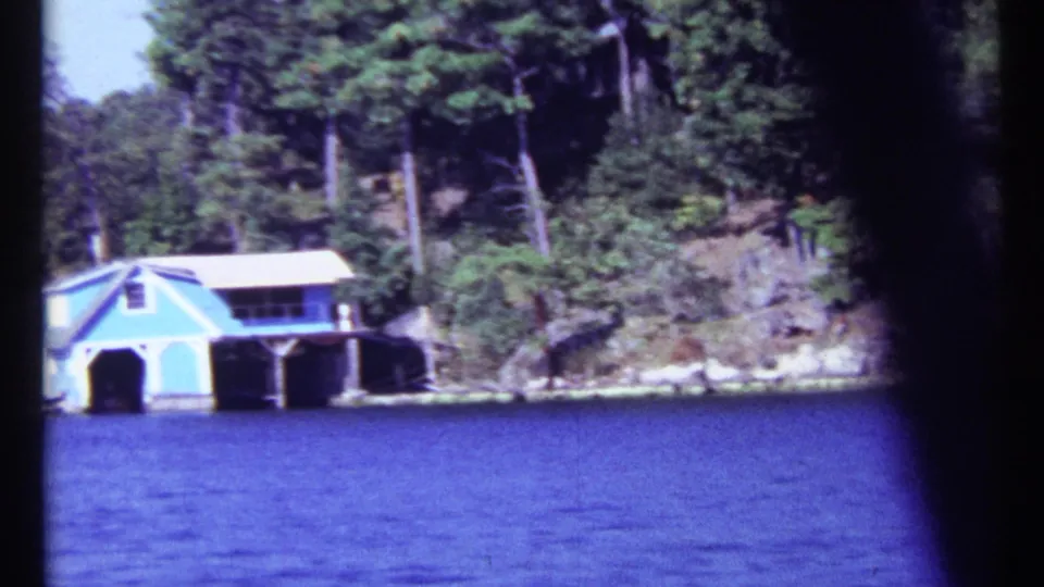 1967 Toronto lakeshore frame showing a blue boathouse near the water, backed by trees, rocks, and shoreline vegetation.