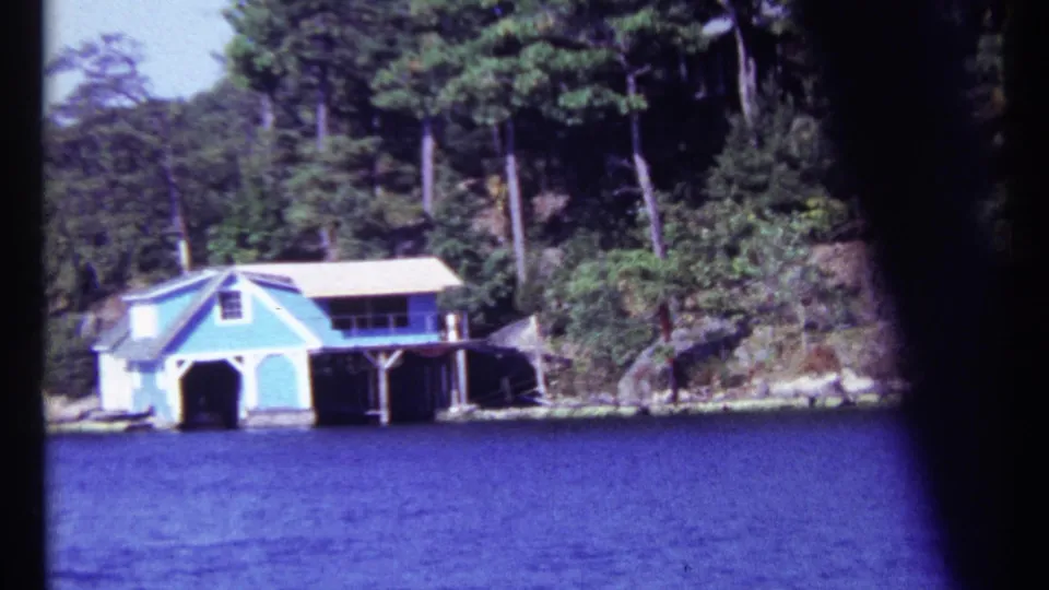 Toronto home-movie frame from 1967 showing a blue boathouse on the lakeshore with dense trees and rocky ground behind it.