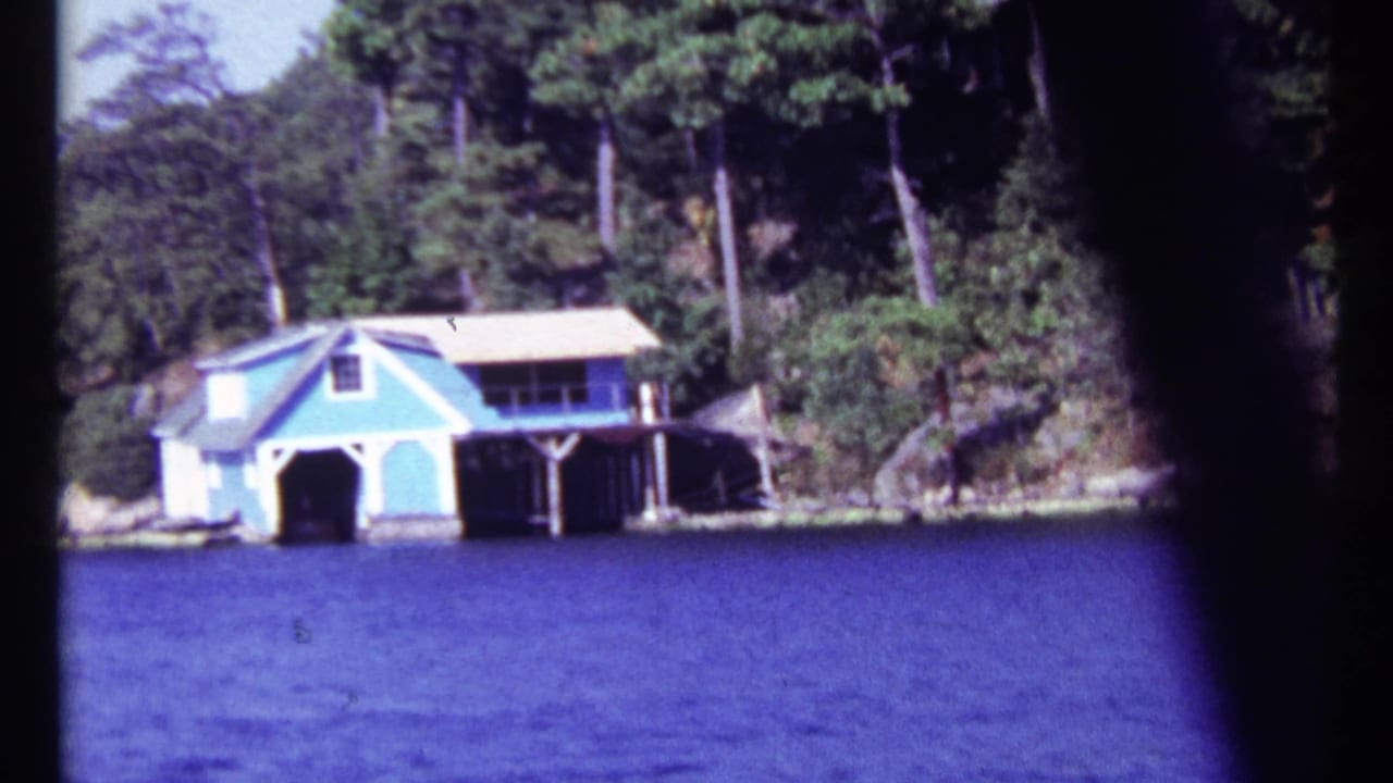 Pale blue boathouse on a wooded rocky shoreline across blue water.
