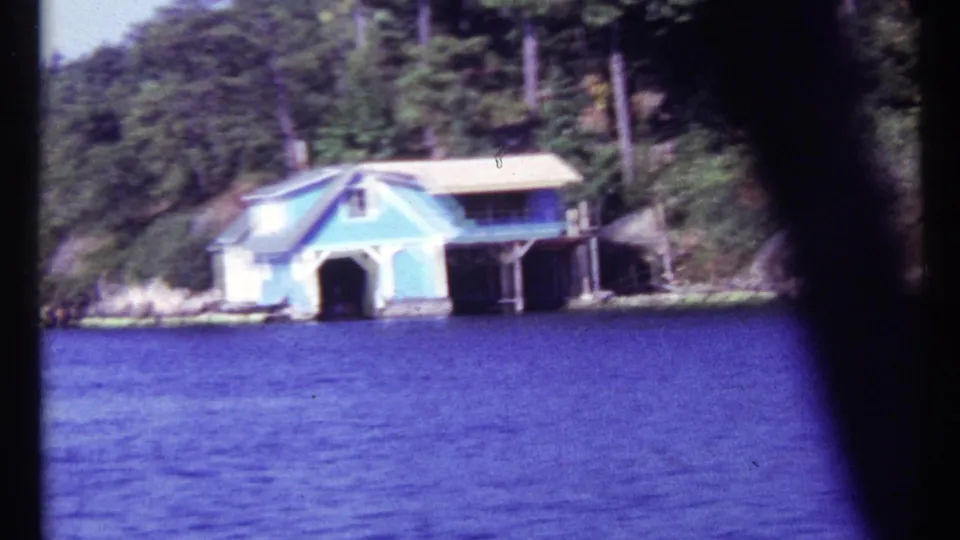 1967 8mm frame from a moving boat showing a pale blue boathouse, dark dock area, rocky bank, and dense trees near Toronto, Canada.