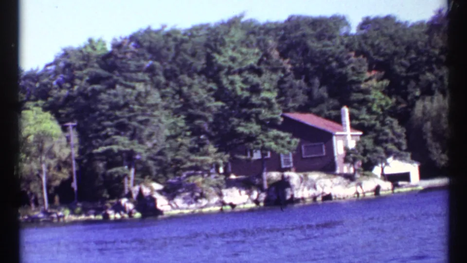 1967 8mm frame showing a brown house with a red roof on a rocky wooded shoreline, filmed from water near Toronto, Canada.