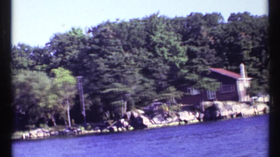 1967 8mm frame from a boat showing a tree-lined rocky shore and a brown lakeside house with a red roof near Toronto, Canada.